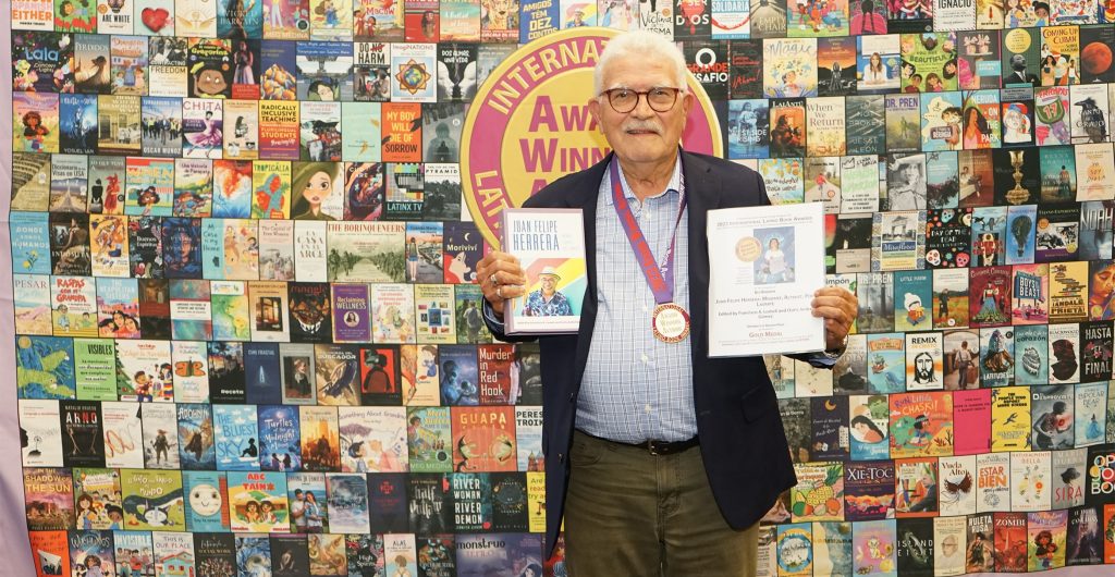 Francisco Lomeli, a smiling man with white hair and glasses, posing beside his book and an award