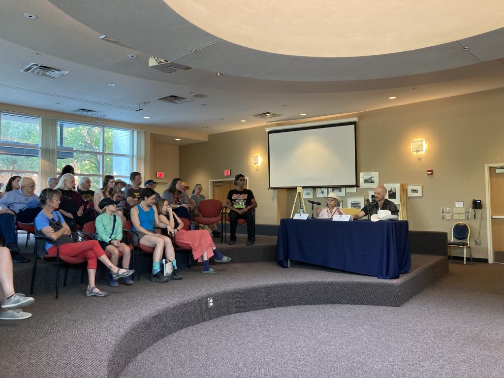 People in a lecture hall listen to two poets sitting at a table and reading from their books.